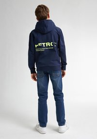 Felpa blu navy con un grande logo giallo sul retro, abbinata a jeans blu e sneakers bianche. Il tessuto sembra liscio e informale.