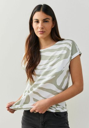 Junge Frau mit langen braunen Haaren, die ein weißes und hellgrünes, zebra-gestreiftes Kurzarmshirt und dunkle Jeans trägt und den Saum des Shirts hält.