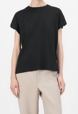 Personne portant un haut noir uni à manches courtes et un pantalon beige, debout devant un fond blanc.