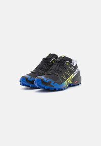 Schwarze Trailrunning-Schuhe mit einer blauen und gelben Sohle. Sie verfügen über ein strukturiertes Obermaterial mit Schnürsenkeln und einer verstärkten Zehenkappe für zusätzliche Langlebigkeit.