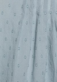 Tessuto blu chiaro con un motivo testurizzato di punti in rilievo distribuiti uniformemente. Il materiale sembra morbido e leggero, adatto per il clima caldo.