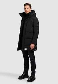Zwarte pufferjas met hoge kraag en capuchon, voorzien van een ritszak op de borst, horizontaal gewatteerd ontwerp en logo-opdruk op de zoom.
