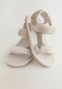 Beige-weiße Sandalen mit mehreren verstellbaren Riemen, strukturierten Sohlen und glatten Akzenten, die ein wellenförmiges Muster auf dem Fußbett aufweisen.