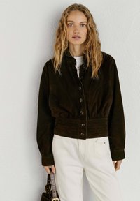 Sieviete ar gariem blondiem matiem, tērpusies tumši brūnā pogājamā jaciņā un baltās biksēs, turot brūnu somiņu pie baltas sienas.