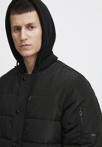 Sort pufferjakke med en tekstureret overflade, som har en lynlåslomme på ærmet og en hætte, der er sikret med snore og trykknapper.