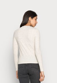 Camisola branca de manga longa com um tricô texturizado, apresentando manchas multicoloridas. Decote redondo e silhueta ajustada, combinada com jeans cinza escuro.