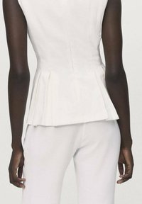 Top bianco a peplo realizzato in tessuto leggero, con scollatura aderente e orlo plissettato. Indossato con pantaloni bianchi abbinati.