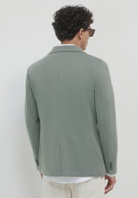 Blazer verde chiaro con tessuto testurizzato, apertura singola e due bottoni sui polsini. Indossato sopra una camicia bianca. Vestibilità rilassata, stile classico.