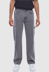 Pantalones cargo grises con bolsillos laterales, cierre de botón y cremallera, y un corte relajado. Llevados con zapatillas blancas sobre un fondo neutro.