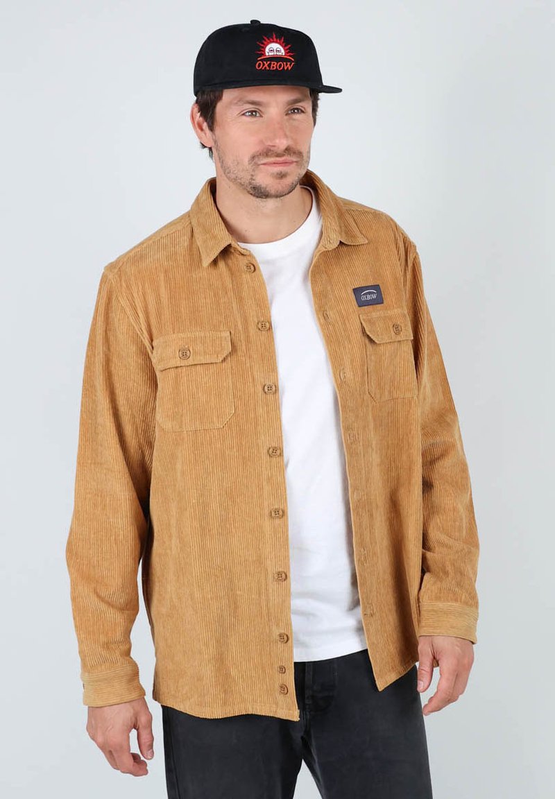 Chemise en velours côtelé couleur sable avec des côtes verticales, présentant une fermeture à boutons, deux poches avant et un patch logo. Portée par-dessus un t-shirt blanc.