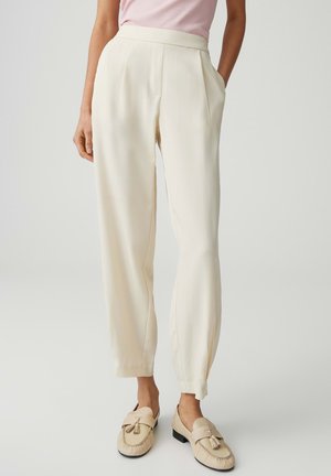 Donna che indossa pantaloni crema a vita alta con tasche laterali abbinati a mocassini crema con nappine e una maglia rosa pallido, in piedi contro uno sfondo chiaro.