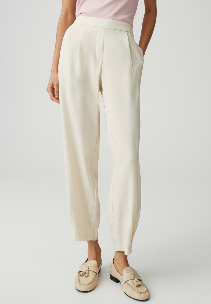 Frau trägt hoch taillierte cremefarbene Hose mit Seitentaschen, kombiniert mit cremefarbenen Quasten-Loafers und einem blassrosa Oberteil, steht vor hellem Hintergrund.