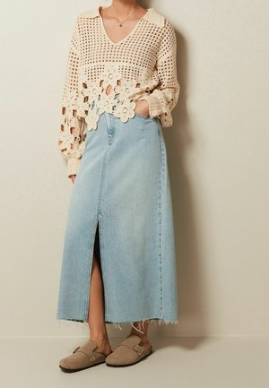 Femme portant un pull en crochet crème, une jupe en jean taille haute bleu clair avec une fente à l'avant, et des chaussures beiges à enfiler avec des boucles.