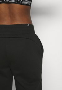 Pantalones deportivos negros con cintura ajustada, bolsillos laterales y un discreto logo de Puma en la parte trasera. Textura suave y diseño de pierna entallada.