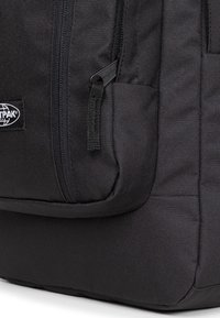 Mochila negra hecha de un tejido duradero con múltiples bolsillos con cierre y una superficie lisa y texturizada. El logo es visible en el lateral.