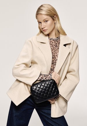 Cappotto oversize beige, top con stampa leopardata, jeans blu scuro. Borsa a tracolla nera trapuntata con manico a catena. Texture dei tessuti liscia e design strutturato.