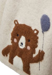 Couverture en laine couleur crème avec un motif d'ours brun tenant un ballon bleu, présentant une texture douce et des détails brodés.