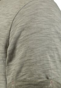 Short-sleeve, olive green shirt made of textured fabric with a subtle pattern. Visible red stitching detail near the hem.