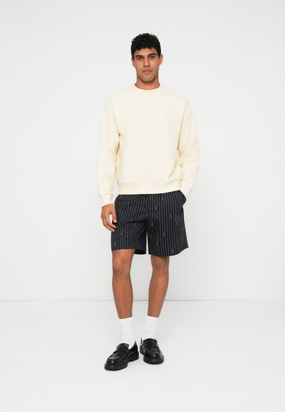 Sweatshirt beige clair, short à rayures noires, chaussettes blanches et chaussures noires. Le sweat a un col ras du cou, le short présente de petits motifs.