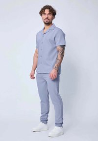 Homme aux cheveux bouclés et aux tatouages sur les bras, portant une chemise à boutons à manches courtes bleu clair et un pantalon assorti, avec des baskets blanches, debout.