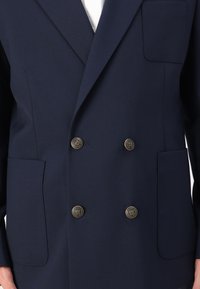 Marineblauer doppelt geknöpfter Blazer aus strukturiertem Stoff, ausgestattet mit vier Taschen und goldfarbenen Knöpfen mit eingeprägtem Logo-Detail.