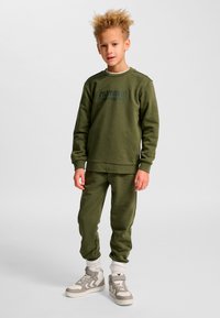 Pojke står iklädd matchande olivgrön sweatshirt och byxor med vita och grå höga sneakers mot en enfärgad bakgrund.