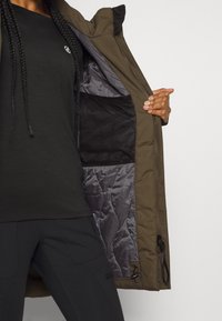 Olivgrön utomhusjacka med grå vadderad foder, meshficka och justerbar dragsko. I kombination med en åtsittande svart topp och byxor.