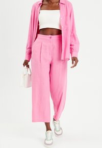 Kvinna iklädd rosa knäppskjorta och byxor, vit crop-top, hållande vit handväska, gående i flerfärgade sneakers på vit bakgrund.