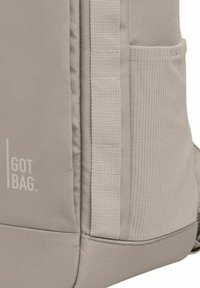 Sac à dos en tissu gris avec une surface texturée, comportant une poche latérale et des accents en sangle verticale. Logo "GOT BAG" affiché.