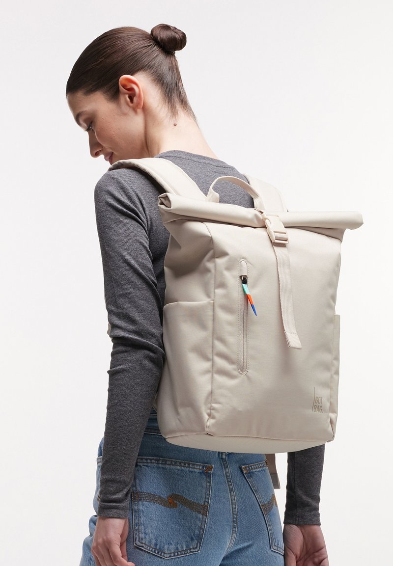Woman with dark hair in a bun wearing a dark gray long sleeve top and blue jeans, carrying a beige roll-top backpack with a colorful zipper pull.
