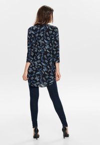 Mörkblå tunika med ett paisleymönster i ljusblått, med trekvartslånga ärmar och en längre bakdel, kombinerad med mörka smala jeans.