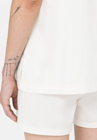 Camicia bianca di cotone con maniche corte, dotata di uno spacco laterale. Abbinata a pantaloni corti bianchi, mostrando un tatuaggio sull'avambraccio scoperto.