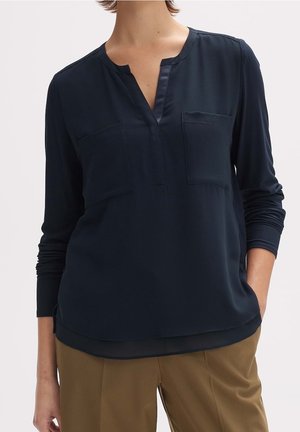 Femme portant un chemisier à manches longues bleu marine avec deux poches poitrine et un col à encoches, associé à un pantalon marron clair.