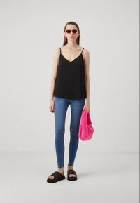 Sort kamiløs top med bølgede kanter, blå skinny jeans, lyserød fuzzy taske, sorte sandaler. Model står mod en ensfarvet baggrund.