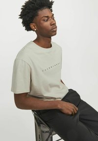 Hellgrau T-Shirt mit schwarzer "JACK&JONES" Schrift, kurze Ärmel, Rundhalsdesign. Das Modell sitzt auf einem schwarzen Netzstuhl.