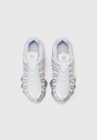 Baskets Nike Shox, avec une tige en maille de couleurs claires, des accents gris supportifs et une semelle distinctive en forme de vague de couleur violette.