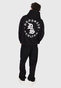 Personne portant un sweat à capuche noir avec le texte "Dropstyle Studios" en blanc dans un design circulaire dans le dos, un pantalon noir et des chaussures blanches.