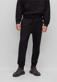 Schwarze Jogginghosen aus weichem Stoff, mit einem elastischen Bund und Kordelzug, mit Bündchen an den Knöcheln und seitlichen Taschen. Getragen mit dunklen Sneakers.