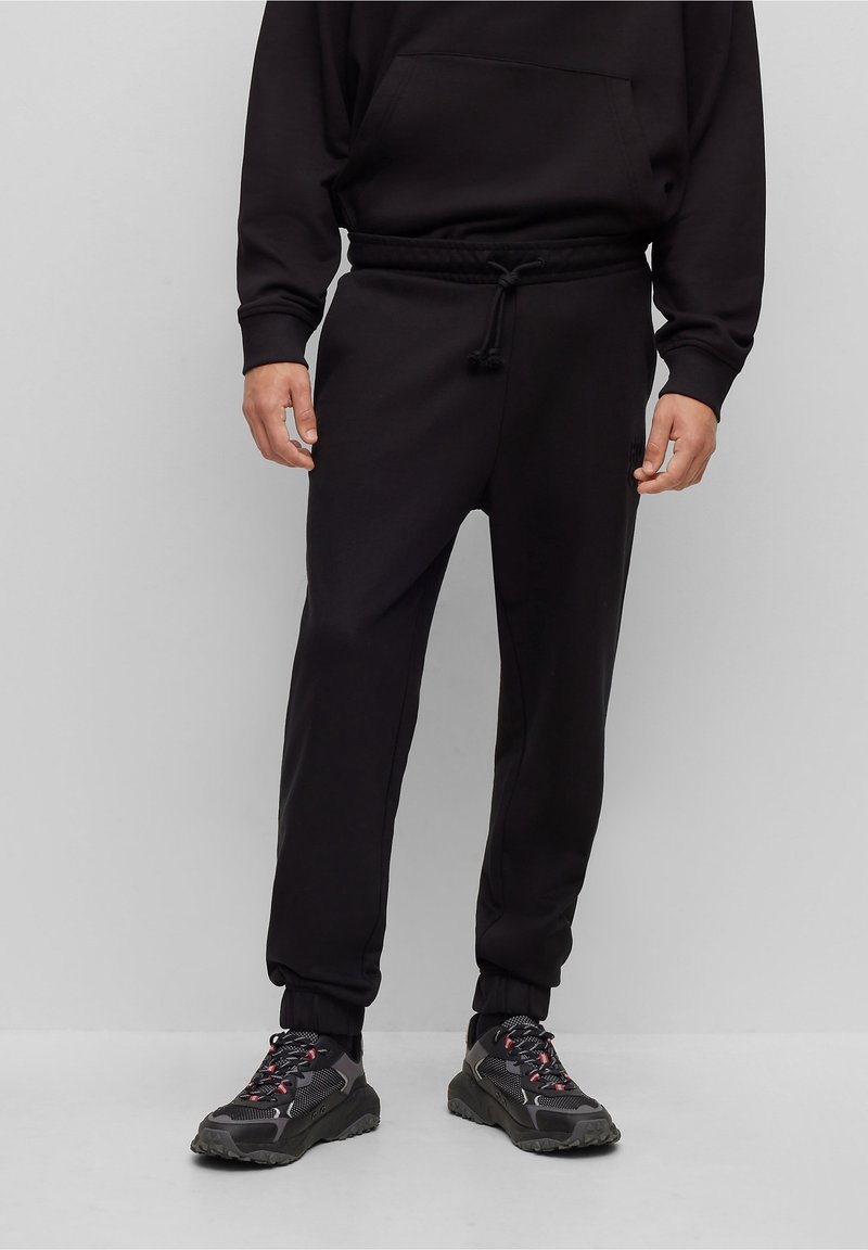 Schwarze Jogginghosen aus weichem Stoff, mit einem elastischen Bund und Kordelzug, mit Bündchen an den Knöcheln und seitlichen Taschen. Getragen mit dunklen Sneakers.