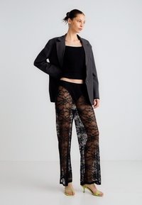 Mustad pitspüksid lillemustriga, kombineeritud istuva musta särgi ja halli bleiseriga. Modell kannab rohelist kontsaga sandaale, positsioneerides end hele taustaga ees.