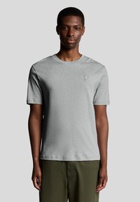 Grå bomulds t-shirt med korte ærmer, rund halsudskæring og lille logo på venstre bryst. Båret med grønne bukser, glat stoftekstur.