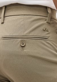 Κοντινό πλάνο στην πίσω τσέπη μπεζ υφασμάτινου παντελονιού με κουμπί και μικρό λογότυπο κοντά στη μέση, φορεμένο με λευκό πουκάμισο που έχει μπει μέσα.