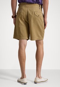 Person som bär tan-färgade shorts med knappar på baksidan, lila skjorta, vita Polo-sneakers, stående mot en plain ljus bakgrund.