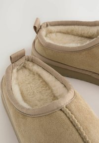 Zapatillas beige con un interior suave y esponjoso y un material exterior liso. Los bordes tienen un ribete reforzado y una lengüeta para facilitar la puesta.