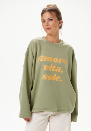 Kobieta o blond włosach związanych w niechlujny kok nosi oversize'ową bluzę w kolorze szałwi zielonej z nadrukiem "Amore, vita, sole." w kolorze żółtym.