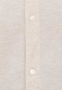 Φωτεινό μπεζ πουκάμισο με κουμπιά, διαθέτει απαλό ύφασμα, τέσσερα λευκά κουμπιά και διακριτικό φυσικό μοτίβο υφάσματος.