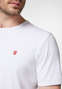 Biała koszulka z krótkim rękawem z czerwonym haftowanym logo na lewej piersi, wykonana z miękkiego materiału bawełnianego, z okrągłym dekoltem.