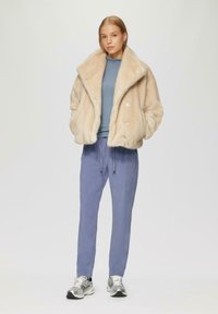 Faux-Fur-Cremüberhemd, hellblaues eng anliegendes Oberteil, lockere lavendelfarbene Hose mit Bundzug. Das Modell trägt silberne Turnschuhe.