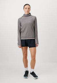 Dlhý rukáv, ľahký šedý športový vrch s vysokým golierom v kombinácii s čiernymi šortkami. Vrch má kontrastné švy a voľný strih.