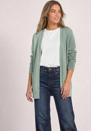 Femme portant un cardigan vert clair sur une chemise blanche et un jean bleu foncé, debout devant un fond clair uni.
